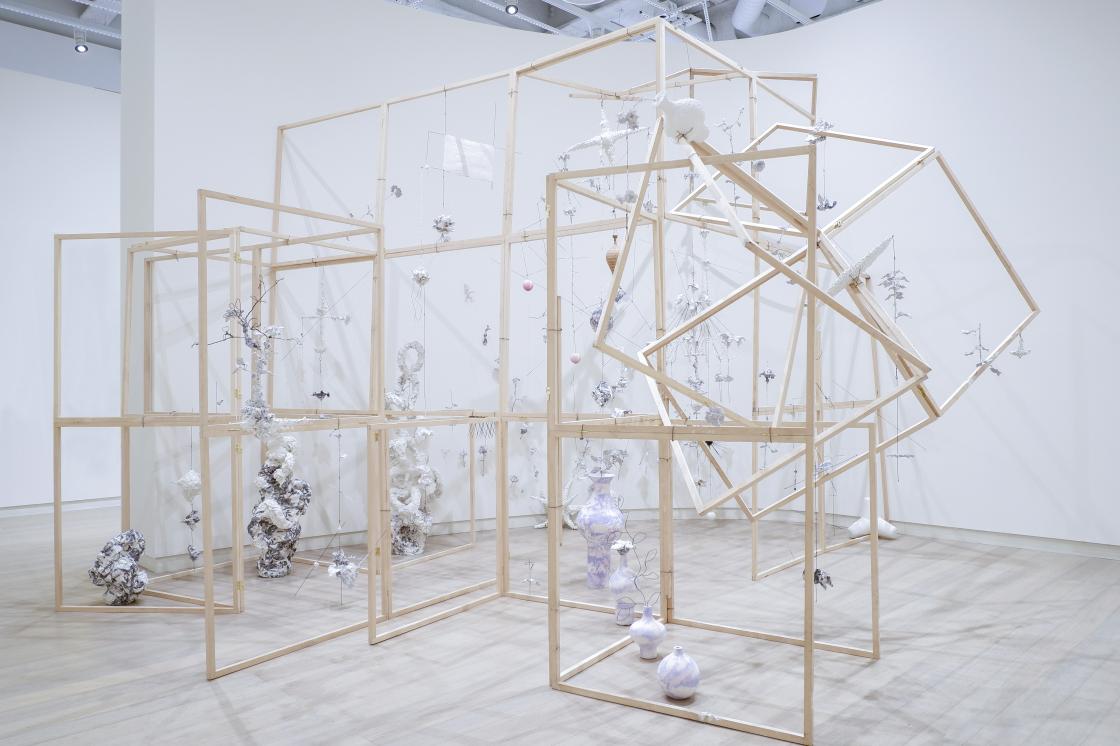 The image is a photo of an installation by the artist Sangmin Lee. The installation is a collection of wooden frames, typically used in construction, positioned within an open gallery space. Each structure is positioned around a series of objects made of delecate materials, from porcelain vases to paper mache sculptures.