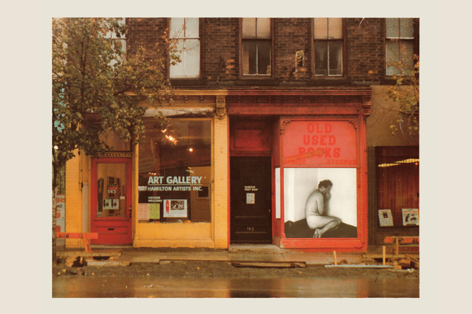 A photo of the streetview of two building fronts, one on the left is yellow and has a sign in the window that reads "art gallery, hamilton artists inc". the other building on the right is red and has a large photo pasted on the front. 