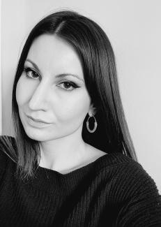The image is a black and white photo of Žana Kozomora. Žana is a while woman with dark hair and is wearing a black top and a hoop earring in her left ear. Her face is tilted to her left and she is smiling and looking directly into the camera.