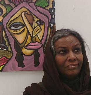 The image is a photo of Saud Badri. Saud is an African woman with dark brown skin and grey hair which is framed by a scarf. She has dark brown eyes and looks off camera while standing in front of a painting.