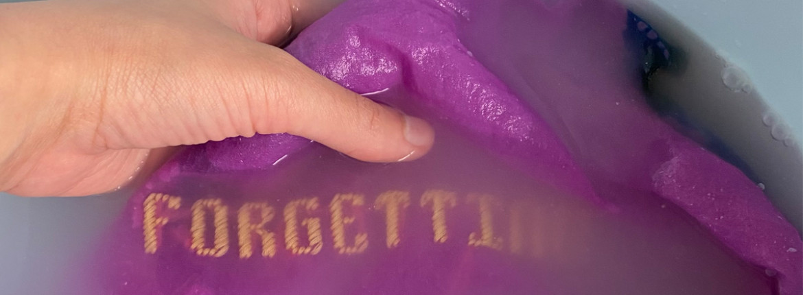 A dyed sheer purple fabric immersed in a cloudy water solution, the word “forgetting” is visible in yellow cross stitch lettering. A hand searches for the word “forgetting” in the cloudy waters.