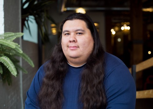 John Hill is looking directly at the camera and smiling slightly. John has long black hair and is wearing a deep blue shirt.