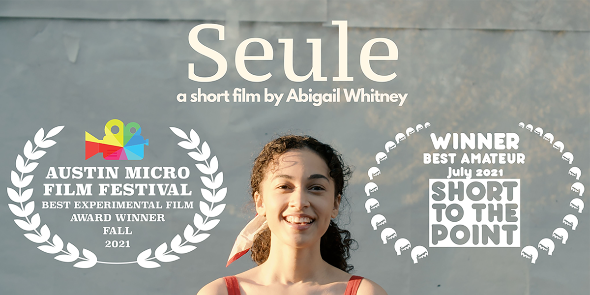 A photograph of the head and shoulders of a young person with long brown curly hair tied back into a low ponytail. They are wearing something with red straps, as well as a red and light pink scarf in their hair. They are smiling brightly. The background is a grey wall. Above the person’s head is the title of the film, “Seule”, and a description that reads, “a short film by Abigail Whitney”. On either side of the person is an award stamp. One reads “Austin Micro Film Festival, Best Experimental Film Award Winner, Fall 2021” and has a rainbow video camera icon. The other reads “Winner, Best Amateur, July 2021, Short to the Point”.