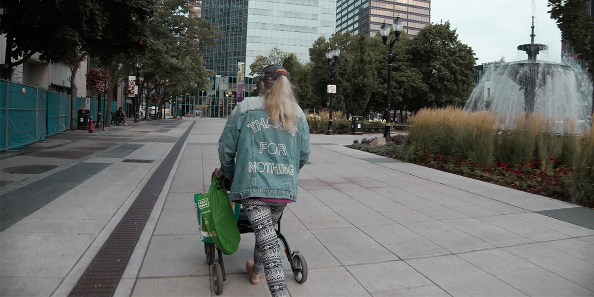 A woman walks down the street. The back of her denim jacket reads “Thanks For Nothing” in white letters. She has light hair and is wearing it pulled through the back of her baseball cap. She is wearing patterned leggings and is pushing a mobility aid. There is a fountain to her right and tall, shiny buildings ahead of her.