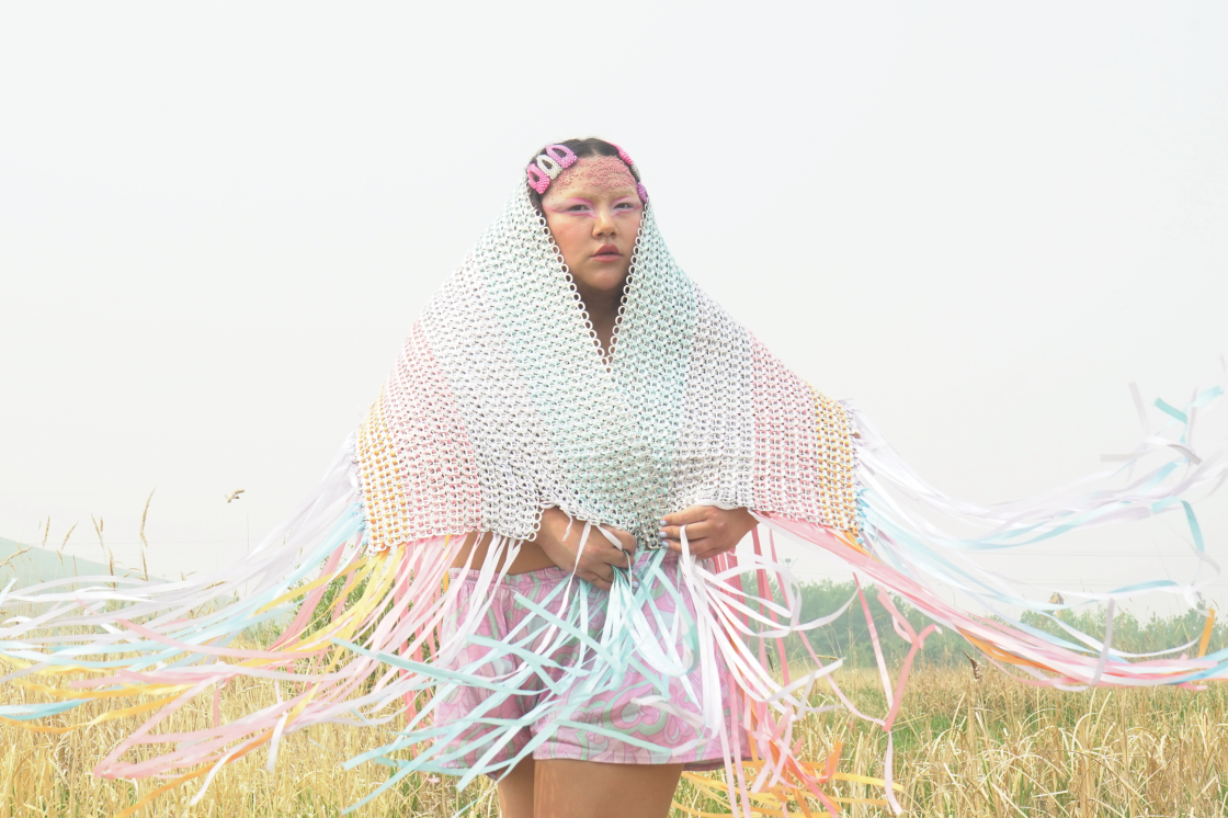 Detail of Cheyenne Rain LeGrande's Billboard photo featuring a subject in a grassy field wearing a pastel coloured Bepsi Tab shawl over their head and covering their shoulders with ribbon fringe along the bottom of the shawl outstretched in the wind