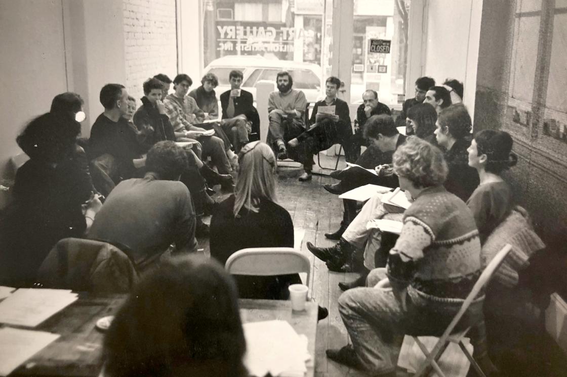 The image is a photo of  Annual general meeting of the Independent Artists Union at Hamilton Artists Inc., Nov. 29, 1987 by Cees van Gemerden. It's a black and white photo which shows a group of artists sitting in a circle on folding chairs in a crowded gallery space.