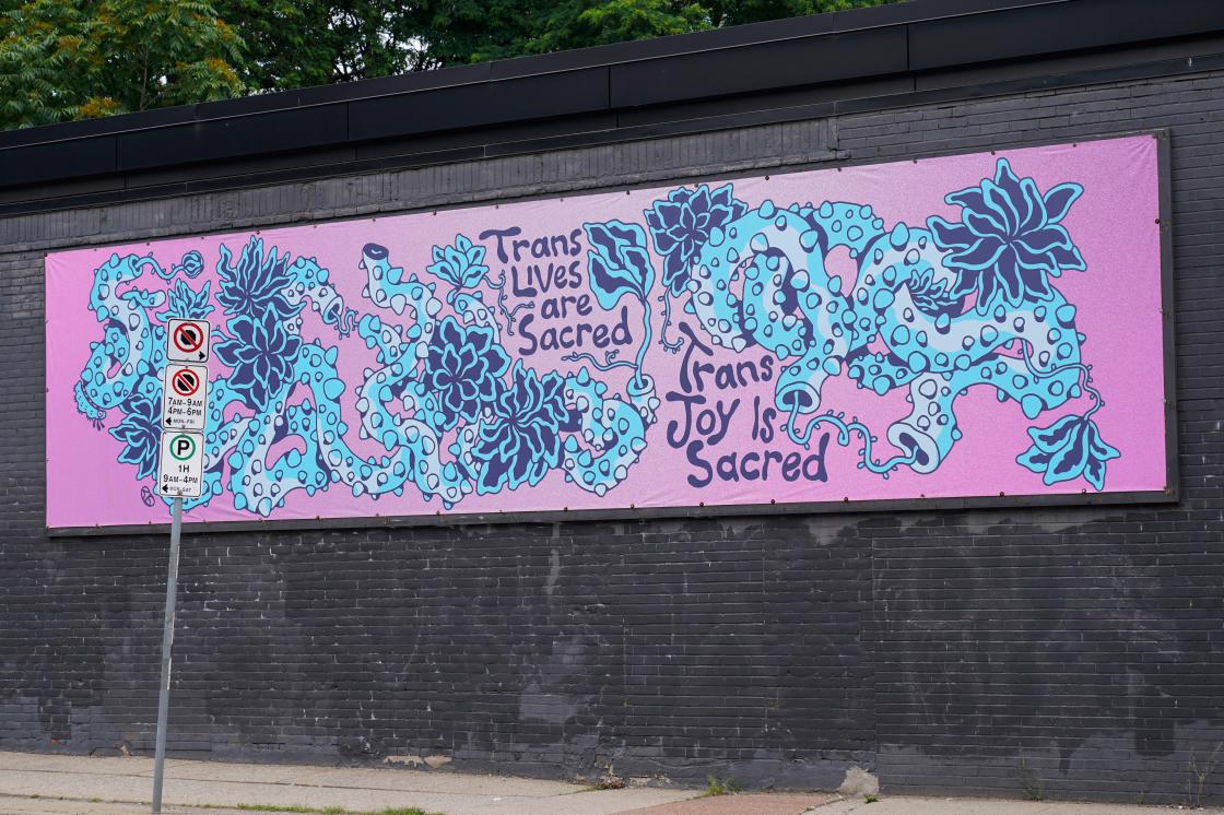 A rectangular purple and blue illustrated banner installed on a gray brick wall on the exterior of Hamilton Artists Inc. The banner is a drawn depiction of blue-coloured vines with thorns intertwined with each other across a wide background of a purple gradient. The vines have light blue highlights and flowers and smaller slender stems are interspersed amongst the coils of the vines. The text “Trans Lives are Sacred'' and “Trans Joy is Sacred” is nestled between the vines in the middle of the illustration.