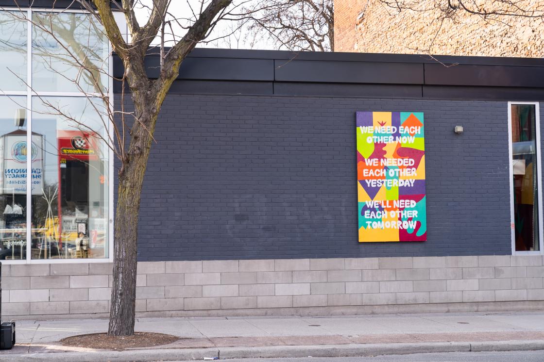 a colourful rectangular mural sits on the external grey brick wall of the Inc's building. The mural has abstract shapes and the text: we need each other now, we needed each other yesterday, we'll need each other tomorrow.