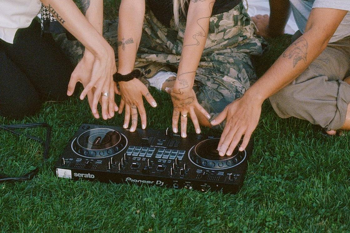 Three pairs of hands touch a CDJ sitting on some green grass.