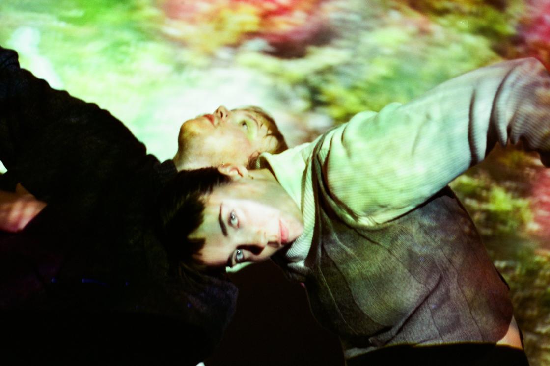 Two people dance in a dark room, illuminated by a green yellow and red abstract projection. They have their heads bent near one another in the centre of the image, their arms flailing outwards.