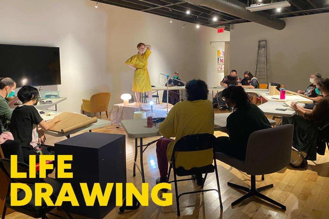 10 people are seated at tables in a circle drawing a model in a yellow robe standing in the center of the tables