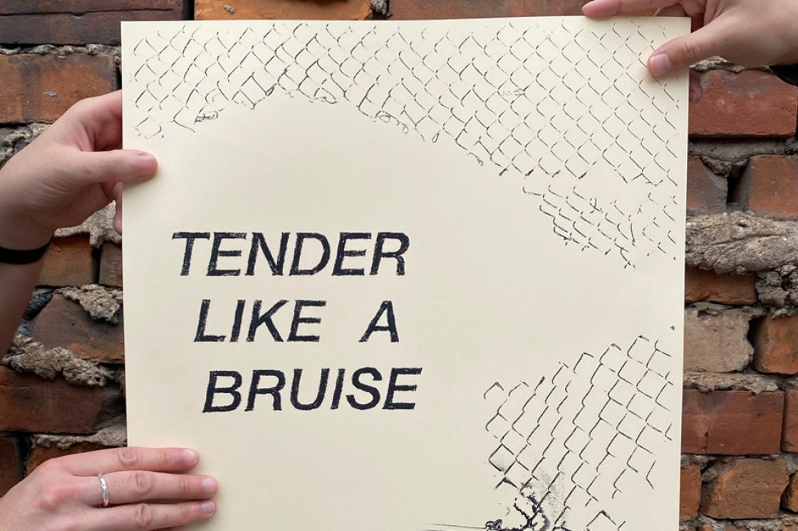 three hands hold a paper beige publication against a red brick wall. The publication reads: TENDER LIKE A BRUISE