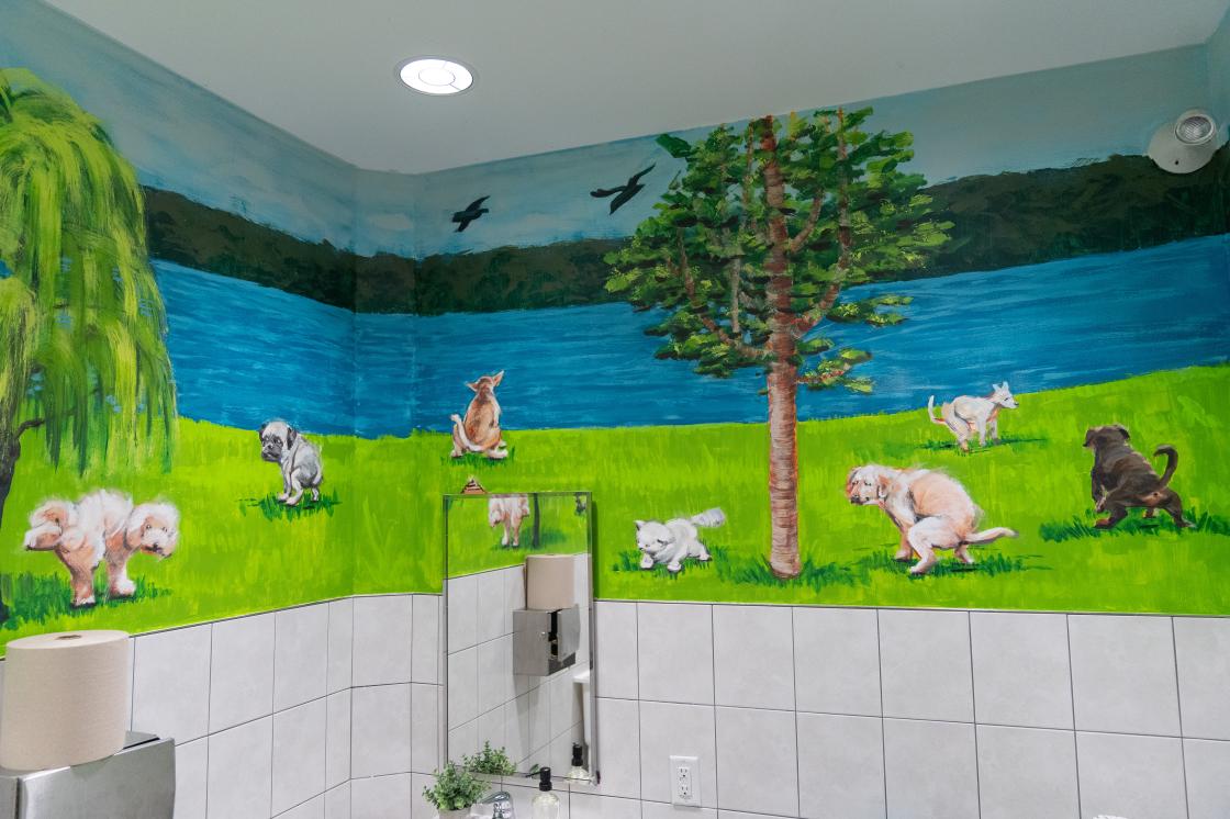 Yigi Chang's mural, "The Call of Nature". The mural is on the top half of the bathroom walls with tile underneath. There are dogs and cats scattered on a bright green grass lawn.