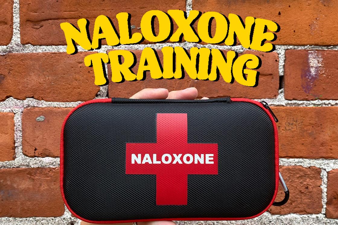 A photo of a red and black Naloxone kit in front of a red brick wall. It says "Naloxone Training" in bold yellow font at the top. 
