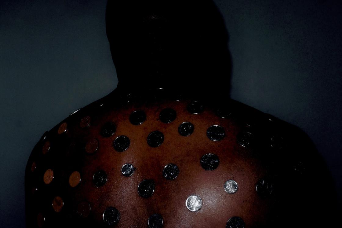 "How much a $$$ cost?" by Deion Squires (2023). A black person is photographed from behind, from their upper back to the top of the back of their head. There's a dim light pointed at the middle-bottom of their right shoulder, and the wall behind them is a dark teal. There are silver coins spaced out evenly, attached to their back, about a couple of centimetres between each coin. 