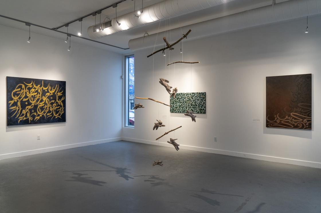 This is a photo of 'Dreams to get you through the night'. There is a mobile featuring pulp-made bunnies floating in the middle of the gallery. One painting by Rania Haider is on the left wall, and two paintings by Rania Haider are on the adjacent wall. There's a window with a blue light casting in.