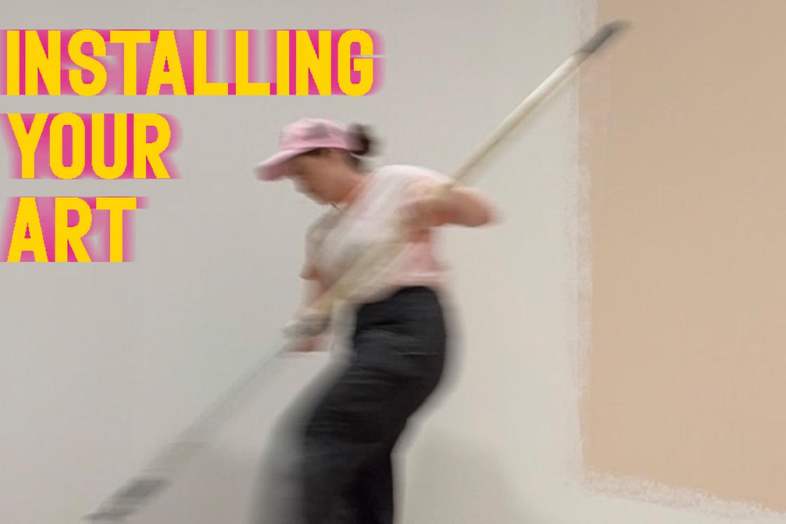 a motion-blurred picture of Bre rolling a long paint-roller in a tray of paint on a gallery floor. She is wearing a pink ball cap. Yellow words: Installing You Art are typed in the top left hand corner, with pink motion blur behind the words. 