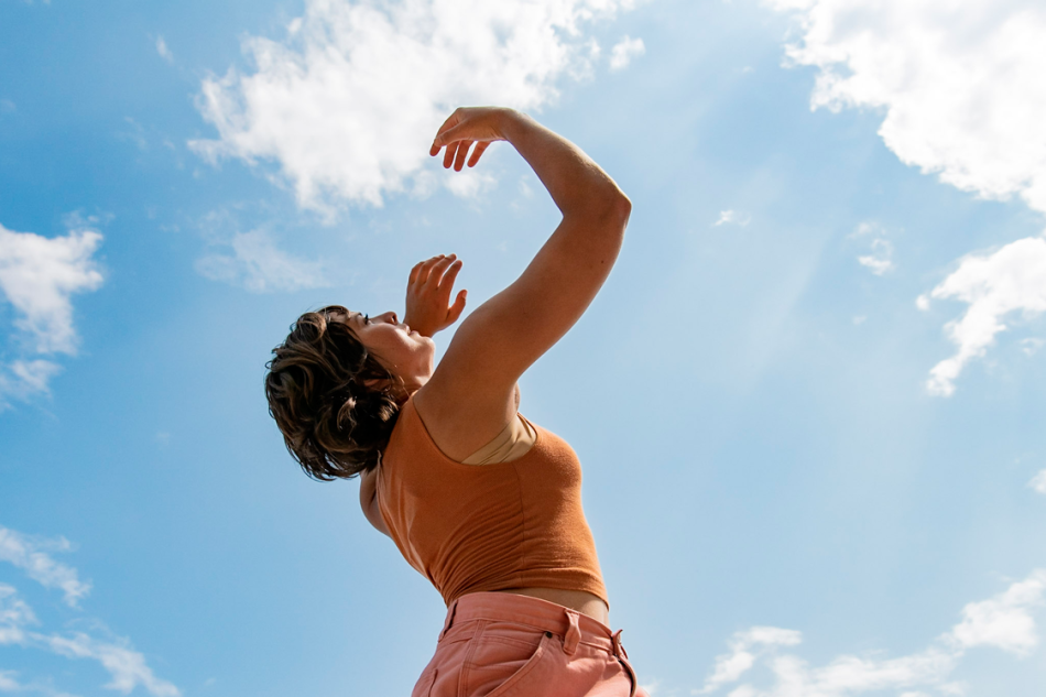 One person stands against a blue sky background with their body facing to the right, their arms in the air