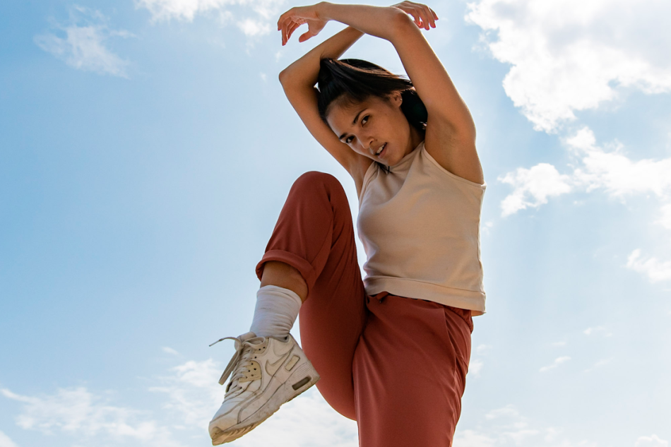 One person stands against a blue sky background, facing forwards, one foot is bent and up in the air with their hands above their head