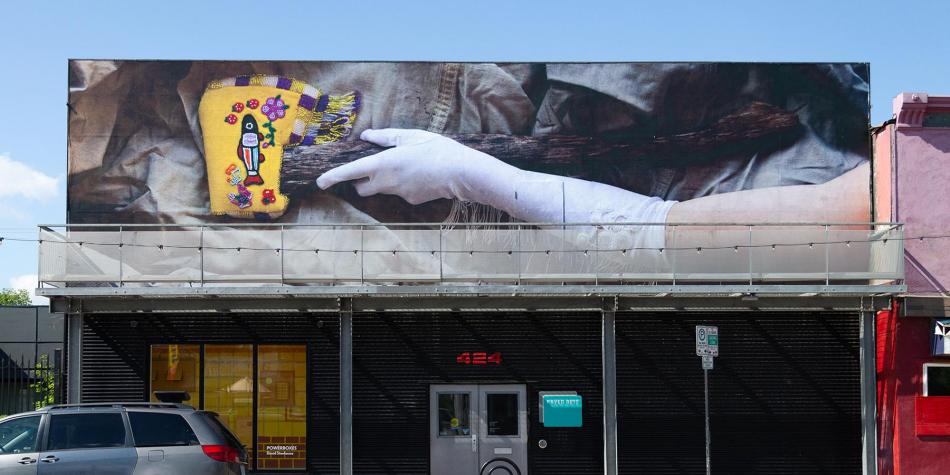 The image is a photo of PAVED Arts. The front of the gallery features a billboard. The billboard features a photograph of a gloved arm holding the handle of an axe. The head of the axe features a sheath that has been decorated with Western North American Indigenous-inspired leatherwork and beadwork.