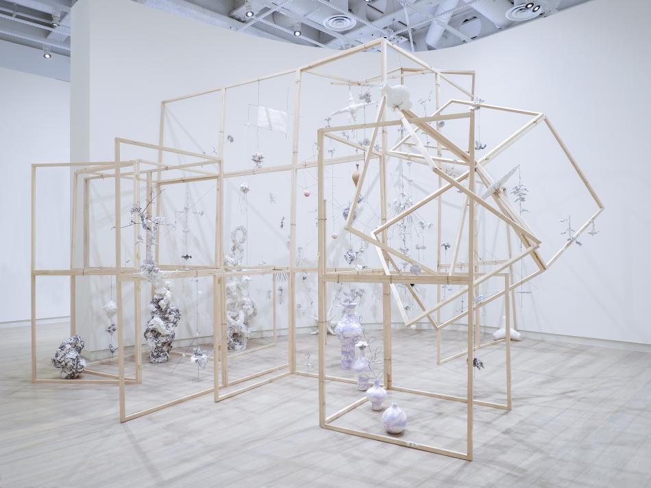 The image is a photo of an installation by the artist Sangmin Lee. The installation is a collection of wooden frames, typically used in construction, positioned within an open gallery space. Each structure is positioned around a series of objects made of delecate materials, from porcelain vases to paper mache sculptures.