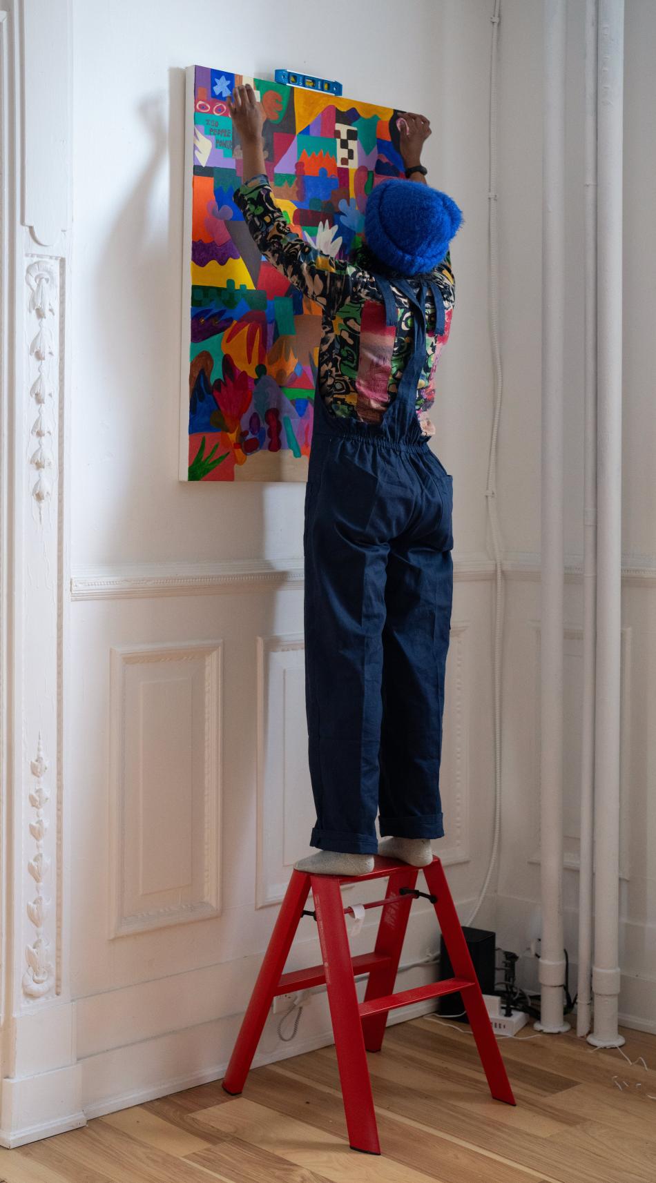 Sahra stands on a red step stool installing their colourful abstract painting on a white wall. 