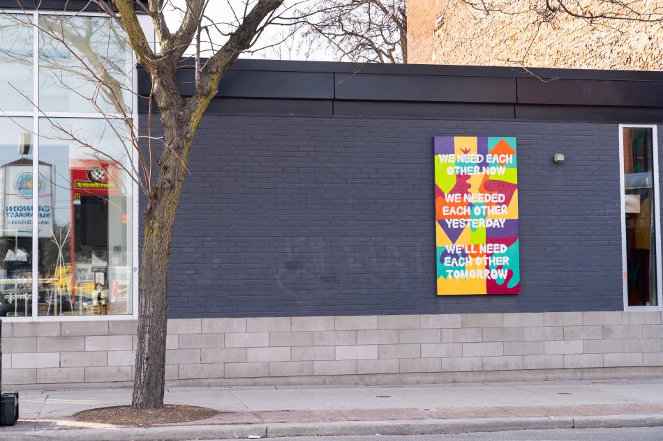 a colourful rectangular mural sits on the external grey brick wall of the Inc's building. The mural has abstract shapes and the text: we need each other now, we needed each other yesterday, we'll need each other tomorrow.