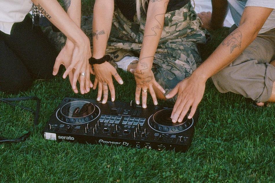 Three pairs of hands touch a CDJ sitting on some green grass.