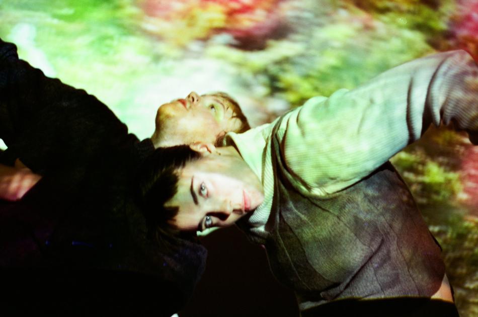 Two people dance in a dark room, illuminated by a green yellow and red abstract projection. They have their heads bent near one another in the centre of the image, their arms flailing outwards.