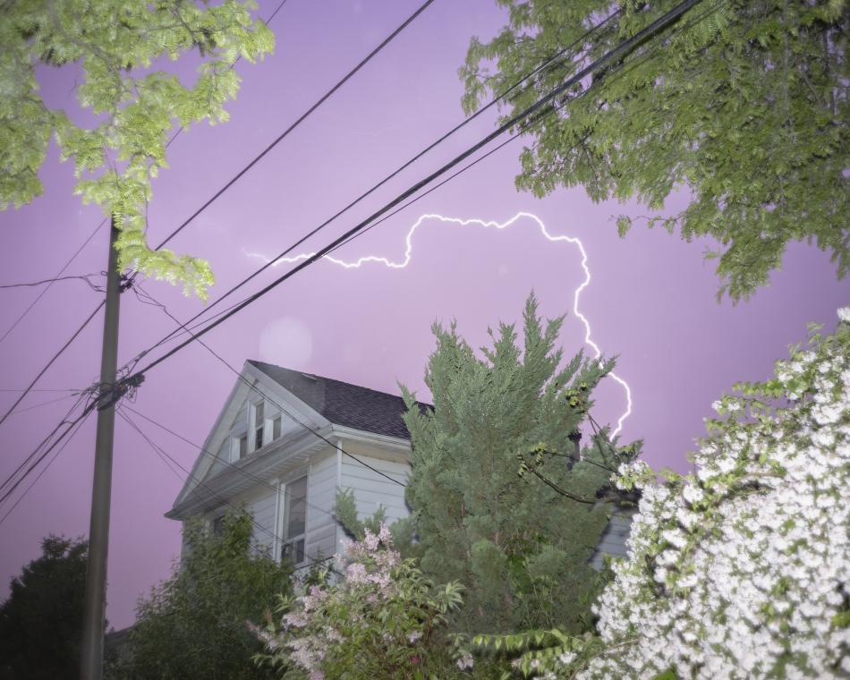 This is Paul Seaton's piece, "Lightning". It features a brick building shot from below, angled upwards, showing a strike of lightning and trees on either side of the photo, and the sky is light purple.