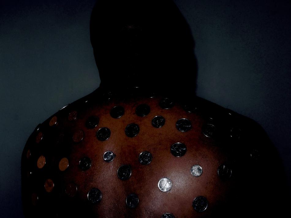 "How much a $$$ cost?" by Deion Squires (2023). A black person is photographed from behind, from their upper back to the top of the back of their head. There's a dim light pointed at the middle-bottom of their right shoulder, and the wall behind them is a dark teal. There are silver coins spaced out evenly, attached to their back, about a couple of centimetres between each coin. 