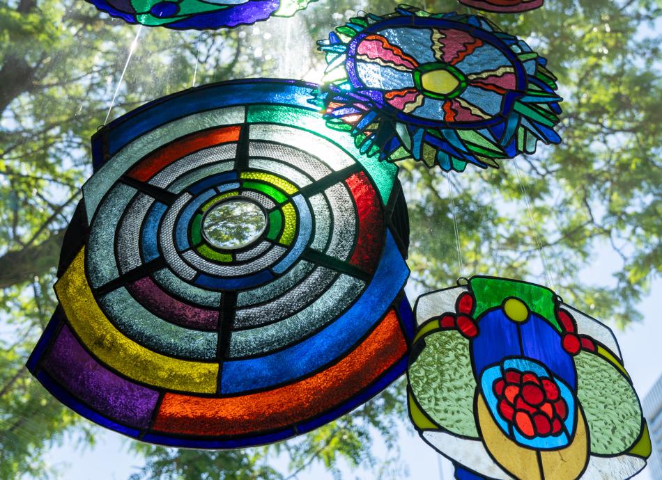 Stained glass works by Mary Porter hanging in a window. The pieces are collaged, asymmetrical, colourful pieces of glass. Trees are behind the window, and light pierces through one of the stained glass works. 