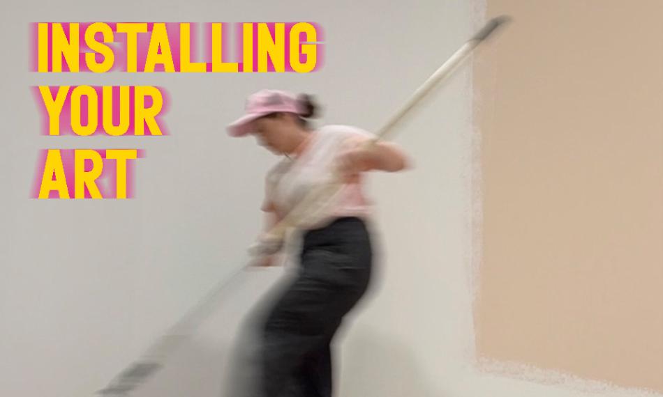 a motion-blurred picture of Bre rolling a long paint-roller in a tray of paint on a gallery floor. She is wearing a pink ball cap. Yellow words: Installing You Art are typed in the top left hand corner, with pink motion blur behind the words. 