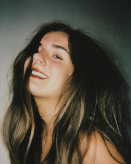 [The image is a photograph of the artist Paige Paton. Paige is a white woman with long brown hair and blue eyes. She is smiling with her face turned upright. Her hair flows freely and partially across her face, obscuring her right eye.]
