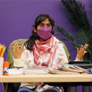 The image is a photo of Sonali Menezes. Sonali is a brown femme with dark hair. She is wearing a purple N95 mask and seated at a table with a lecture microphone in front of her. In front of her, on the table, is a number of materials for making zines; paper, pencils, scissors, etc. Behind her is a dark purple wall and green plants.