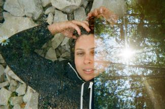 A multiple-exposure film image of Mikah Fortier. They have brown hair and white skin, and are wearing a black hoodie with their arms above their head. Overlaid is an image of trees with sunlight poking through.