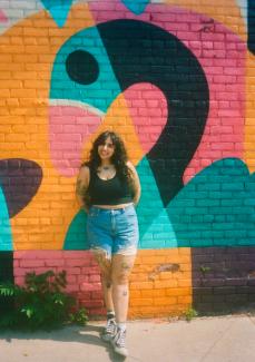 A woman with long black hair wearing a black tank top, black converse and denim shorts stands with her back against a colourful painted brick mural and smiles at the camera