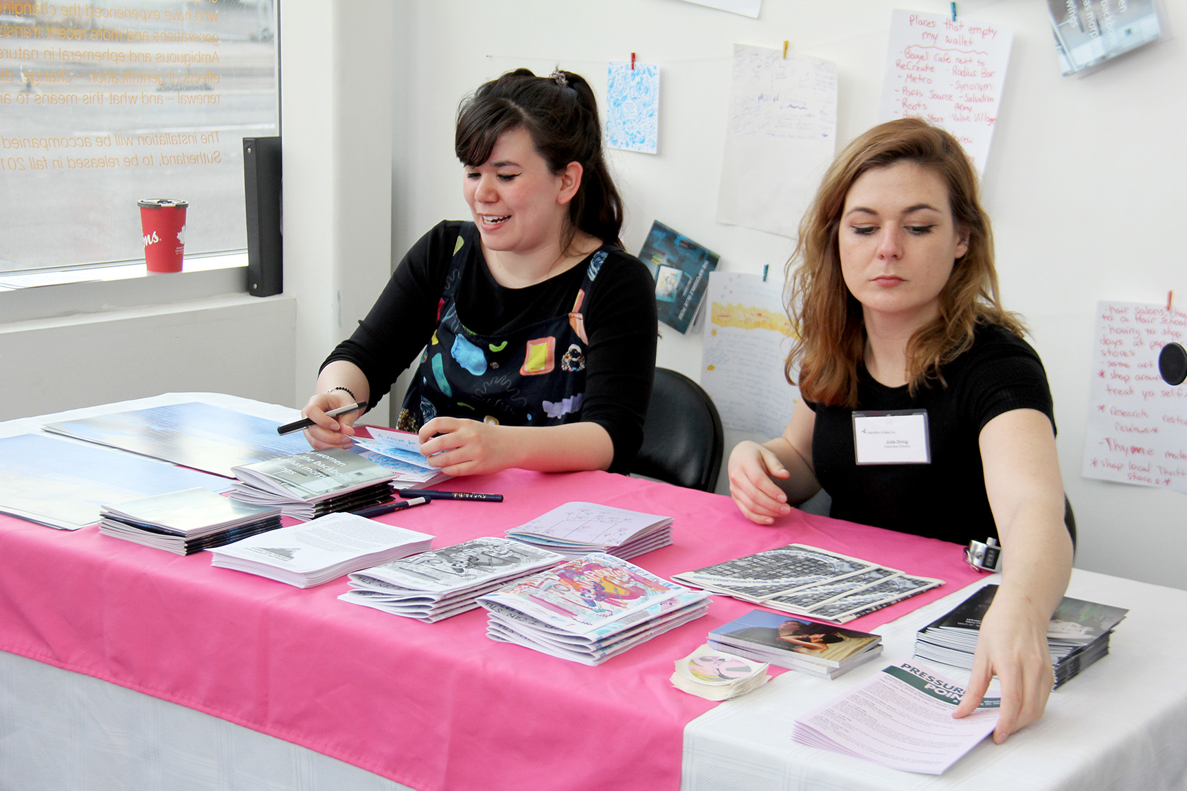 A brightly lit photo of two people seated behind the Zine and Reading corner, which was placed right behind the Inc.’s front door. One person is Julie Dring, a white woman with mid-length wavy hair and winged eyeliner. She is reaching over an assortment of zines and other printed materials on the table in front of her.