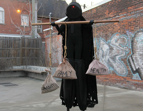 The hooded figure holding the wooden rod, which has three burlap bags hanging from it. The figure looks as if it is offering them to you. Each bag has a label on it: one says “community,” one says, “family,” and the last says “health.” The “community” bag is hanging slightly lower than the others.
