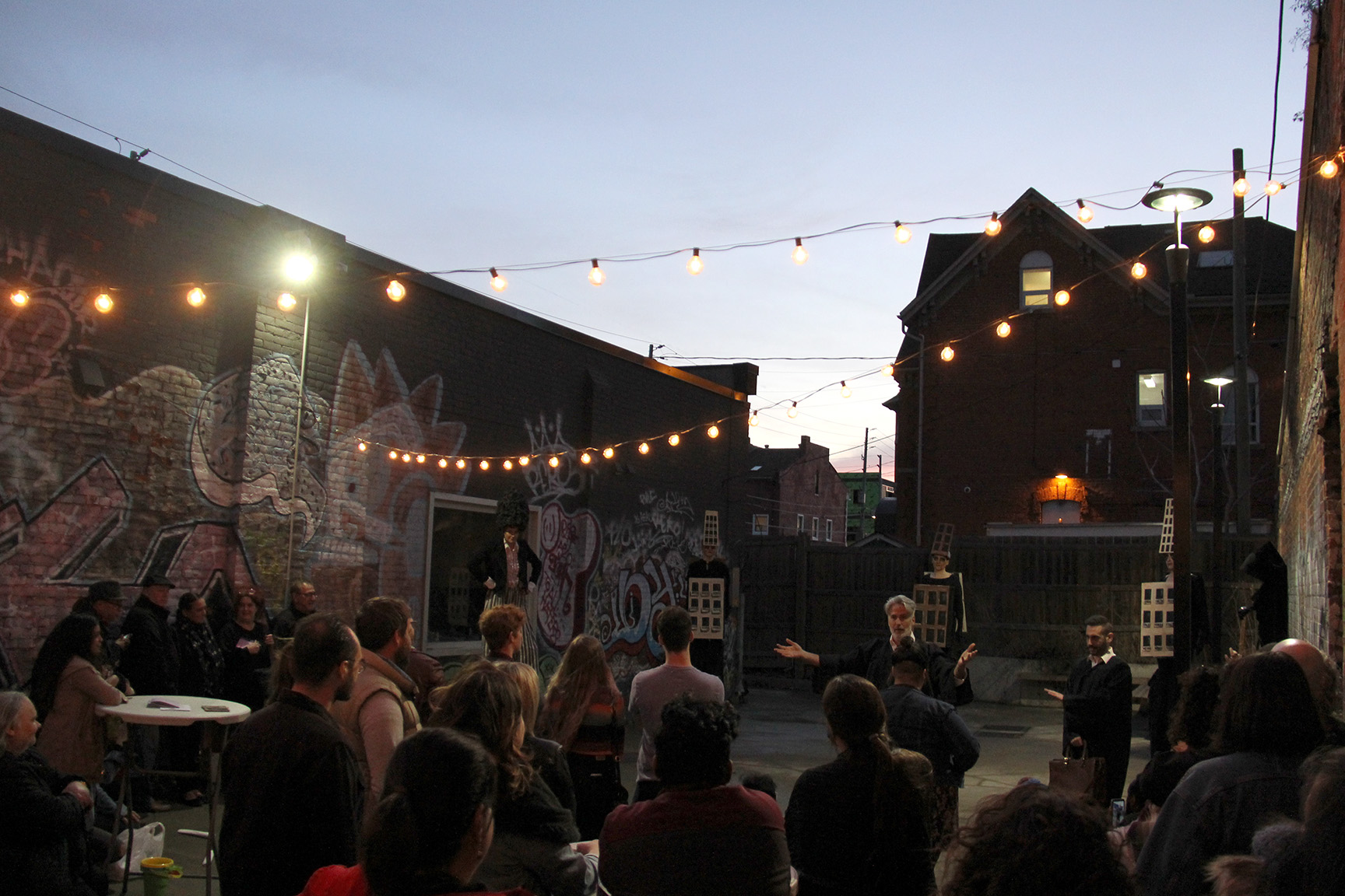 A photo of the Inc.’s courtyard at dusk, framed by the silhouette of the Notre Dame House in the background. There are strings of warm, spherical lights attached to the brick walls on either side of the courtyard. There are 20 or so people seated on fold-out chairs, watching a performance. The performers are dressed in black robes.