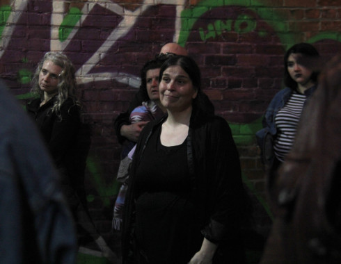 One of the performers, who appears to be a white woman with dark, straight hair that is pulled back. She looks like she has a smug grin on her face. In the background, there are some people standing who appear to be in the audience. 