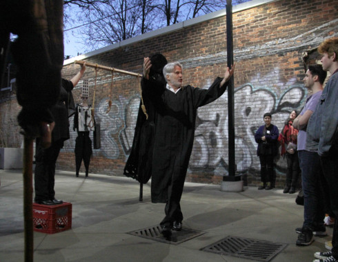 The performer with silver hair and a beard, who appears to be a white man, with his arms raised, as if addressing the audience. He stands in front of the other performers, and several audience members stand just in front of him.