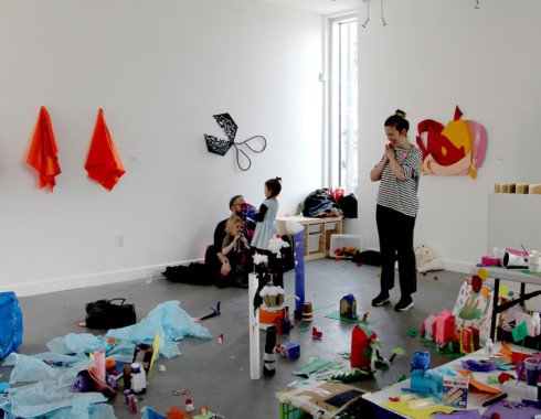 A photo taken from the top of the miniature city. A person in a striped shirt is standing beyond the tissue paper lake. They are smiling with their hands clasped against their chest. There is another adult sitting on the floor with two children.