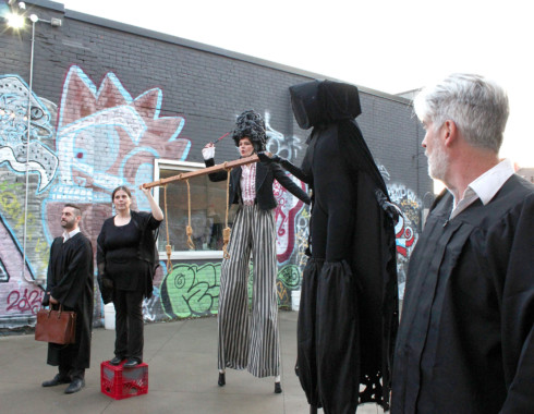Five performers dressed in black standing in a row in the courtyard. From left to right there is: a person in a black robe holding a briefcase, a person holding one end of a wooden rod with three rope nooses hanging from it, a person standing above with stilts and long striped pants, a hooded figure holding the other end of the rod, and a person with silver hair and a beard. 