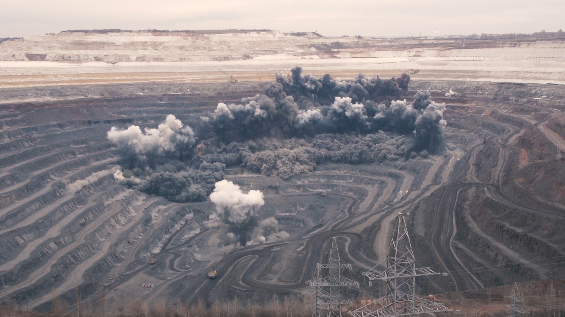 A still from the film "Extractions" by Thirza Cuthand. The still features a mining blast, shot from an aerial view. Large plumes of black smoke dot the landscape, with construction and mining equipment littered about the scene.